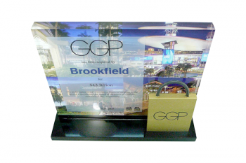 A gold-painted plexi shopping bag in front of a Lucite panel with images on back and transaction details on a frosted acetate.